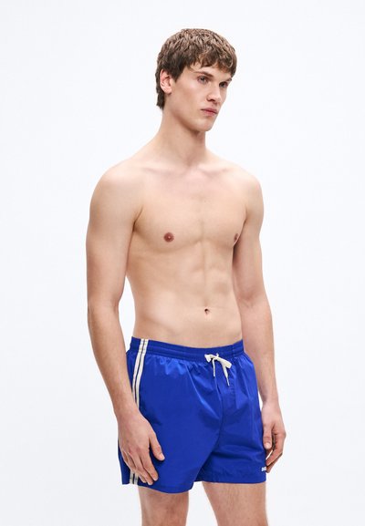Pantaloni scurți de baie albaștri cu dungi albe pe laterale, talie elastica și șnur. Confecționați din material lucios și ușor. Lungime până la mijlocul coapsei.