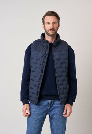 Homme barbu portant une veste matelassée bleu marine par-dessus un pull foncé et un jean bleu, debout devant un fond clair uni.