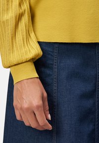 Manche de pull rayé jaune avec un poignet serré, associée à une jupe en denim sombre, mettant en valeur une texture lisse et des couleurs contrastées.