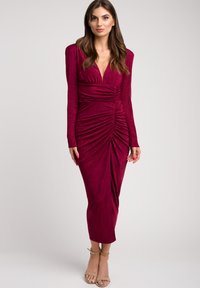 Długa burgundowa sukienka z długim rękawem, głębokim dekoltem w kształcie litery V, marszczeniami i fakturowanym materiałem, zwężająca się do długości połowy łydki.