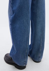 Τζιν παντελόνι με φαρδύ πόδι σε μεσαίο μπλε χρώμα, με ελαφρώς ξεφτισμένο τελείωμα και αντίθετη ραφή, συνδυασμένο με μαύρα δερμάτινα μοκασίνια.