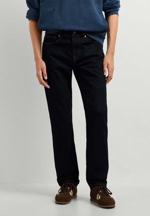 Persoon die donkere blauwe jeans, bruine sneakers en een blauwe trui draagt, staand tegen een effen lichte achtergrond.
