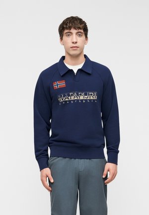 Mladý muž v tmavomodrej mikine s nášivkou nórskej vlajky a vyblednutým logom "Napapijri geographic", stojaci pred jednoduchým pozadím.