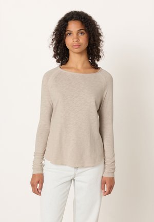 Young woman with curly hair wearing a beige long-sleeve top and light-colored jeans, standing against a plain off-white background.