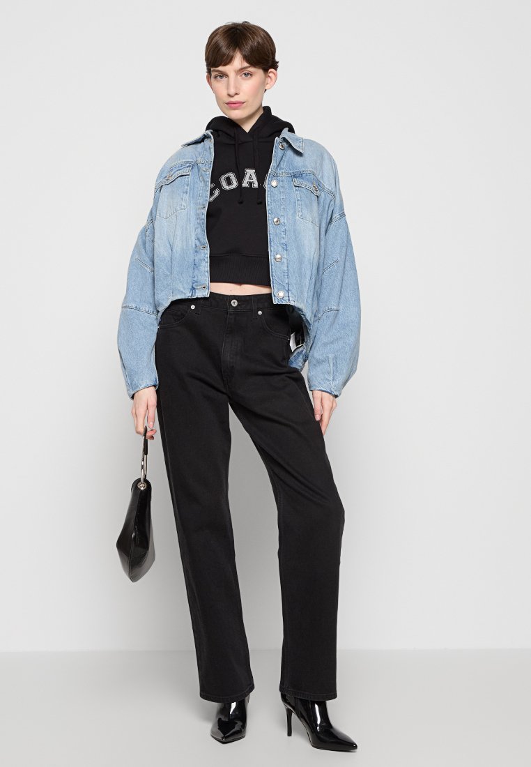 Frau in einer kurz geschnittenen Jeansjacke über einem schwarzen Hoodie mit Aufdruck, schwarzen hochtaillierten Jeans, schwarzen spitzen High-Heels und mit einer kleinen schwarzen Tasche.