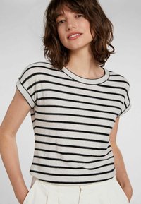 Femme aux cheveux bruns mi-longs portant un t-shirt à manches courtes blanc et noir à rayures horizontales et un pantalon de couleur claire.
