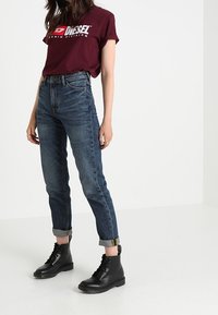 Burgundy bomulls-t-shirt med vit logotyp, högmidjade blå denimjeans och svarta läderkängor med snörning. Jeansen är uppvikta vid anklarna.
