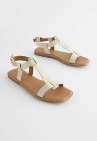 Två vita läder-sandaler med platt sula, med korslagda remmar, ankelspänne och brun innersula. Enkel design med sömnadsdetaljer.