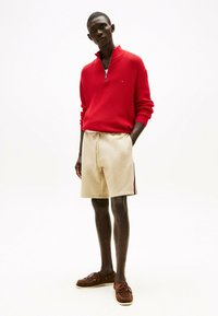 Maglione rosso a zip, pantaloni corti beige con coulisse e strisce laterali, e mocassini marroni. Un abbigliamento semplice e casual con un netto contrasto di colori.