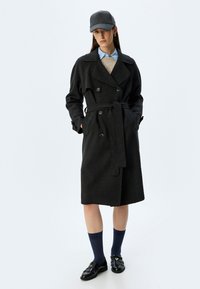 Donkergrijze wollen trenchcoat met een dubbelbreasted ontwerp, brede revers, riem en zijzakken, gedragen over een beige trui en een blauwe blouse.