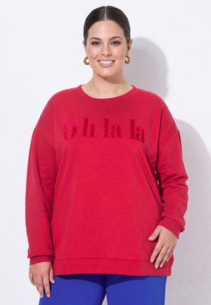 Lächelnde Frau mit goldenen Creolen trägt ein rotes Sweatshirt mit der Aufschrift "Oh la la" und blaue Hosen, stehend vor weißem Hintergrund.
