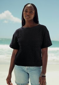 Sort, strikket top med et geometrisk blondermønster, brede korte ærmer og en lige kant, parret med lyse denimshorts.