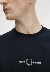 T-shirt nera in cotone con collo girocollo, caratterizzata da un testo ricamato bianco "FRED PERRY" e un design di alloro sul lato sinistro del petto.