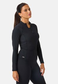 Svart idrottsjacka med en åtsittande design, ribbat mönster, hög krage och fullständig dragkedja fram. Påfallande gulddetalj nära fållen.