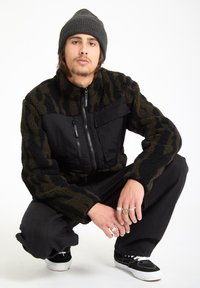 Musta ja oliivinvihreä fleece-takki, jossa on vetoketjulla varustettu etuosaa, teksturoitua kangasta ja rintatasku. Yhdistetty mustiin housuihin ja lenkkareihin.
