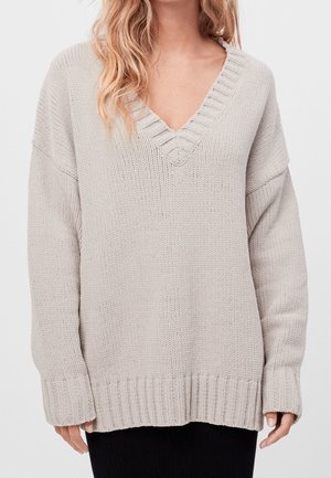 Vrouw draagt een oversized lichtgrijze gebreide V-hals trui met geribde manchetten en zoom, gecombineerd met een zwarte rok.