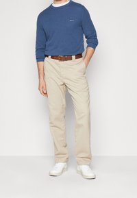 Homme portant un pull bleu à manches longues, un pantalon beige avec une ceinture marron, et des baskets blanches, debout, une main dans la poche, contre un mur blanc.