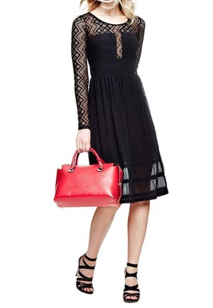 Femme vêtue d'une robe noire à manches longues en dentelle et de talons noirs à lanières, tenant un grand sac à main rouge par ses anses.