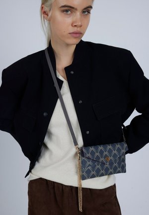 Šedá kabelka cez rameno s vzorovaným dizajnom, zlatým retiazkovým popruhom a modrými prvkami. Nosená cez béžový sveter a čiernu bundu.