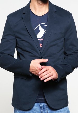 Mand iført navy blazer over grafisk navy T-shirt og lyseblå jeans, hænder samlet ved taljen mod en ensfarvet baggrund.