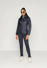 Marineblauwe pufferjack met horizontaal gestikt patroon, voorzien van een rits aan de voorkant en zijpanelen. Gekleed met donkere broek en beige schoenen.