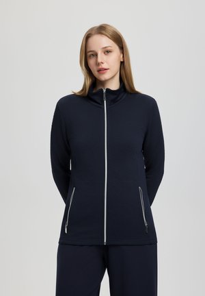 Frau mit glattem blondem Haar, die eine marineblaue Jacke mit Reißverschlusstaschen und weiß umrandeten Reißverschlüssen trägt, vor neutralem grauem Hintergrund steht.