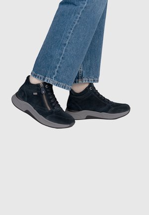 Pair of black suede sneakers with side zipper worn with beige socks and blue denim jeans against a plain light background.