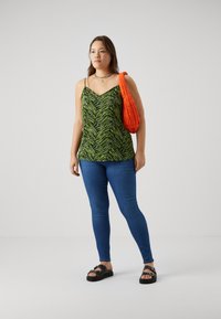Camiseta sin mangas de patrón verde y negro con tirantes finos, combinada con jeans ajustados azul y sandalias negras con tachuelas decorativas. Bolsa naranja.