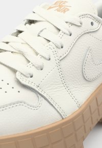 Hvide lædersneakers med en struktureret overdel, perforeret tåboks og en lys brun gummisål med et gittermønster. "NIKE AIR" logo er til stede.