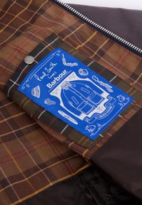 Barna kockás szövet kék címkével. A címke illusztrációkat és szöveget tartalmaz, beleértve a "Paul Smith szereti a Barbourt" feliratot. A cipzár részlet látható.