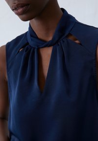 Granatowa bezrękawnikowa bluzka z gładkiego materiału o wysokim dekolcie, z kluczem z przodu i plisowanymi detalami na ramionach.