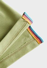 Grøn stof med en struktureret vævning har farverige stribede accenter ved kanterne i blå, gul og orange, der viser syede sømme.