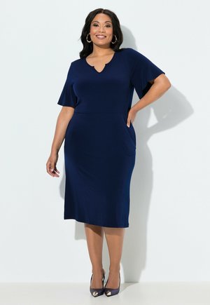 Uśmiechnięta kobieta stoi z ręką na biodrze, ubrana w granatową sukienkę do kolan z falbanowymi rękawkami oraz czarne szpilki z złotymi czubkami.