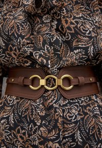 Ceinture en cuir marron avec une boucle circulaire dorée, resserre un tissu à motif floral noir et marron. Texture lisse, sangle réglable.