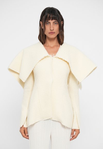 Cream textured jacket with wide, layered collar and long sleeves. Features a front closure and flared hem. Worn with light-colored pants.