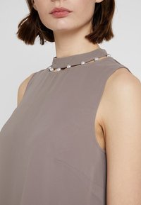 Blouse sans manches couleur taupe avec un col montant, ornée d'un détail en perle et confectionnée dans un tissu léger et lisse avec un léger brillant.