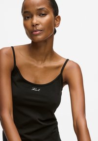 Melnā bezpaliktņa blūze ar plānām siksnām, ar izliekta kakla izgriezumu un nelielu balto logotipu izšuvumu priekšpusē. Gluda auduma tekstūra.