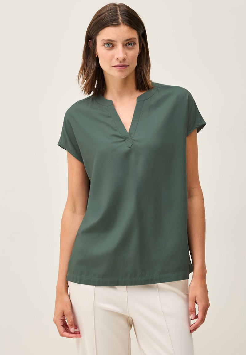 Blusa verde a maniche corte con scollatura a V, caratterizzata da una texture liscia e una vestibilità rilassata. Abbinata a pantaloni color crema.
