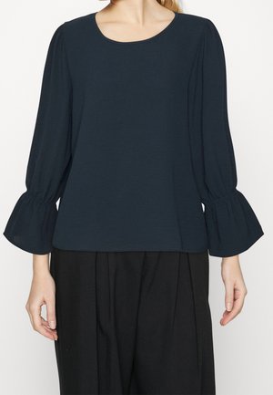 Femme portant un chemisier bleu marine foncé avec des manches cloche froncées et un col rond, associé à un pantalon noir ample.