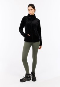 Schwarze, Reißverschluss-Fleecejacke mit hohem Kragen und zwei Seitentaschen, kombiniert mit olivgrünen gerippten Leggings und schwarzen Wanderschuhen.
