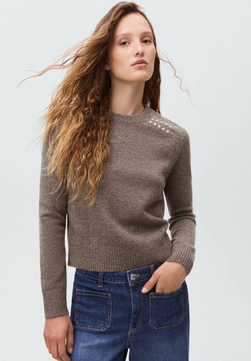 Brauner Strickpullover mit geripptem Saum und Bündchen, verziert mit dekorativen Knöpfen an der linken Schulter; kombiniert mit blauen Jeans.