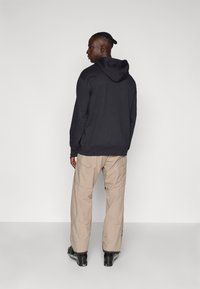Zwarte hoodie met een ontspannen pasvorm, gecombineerd met beige cargobroek met meerdere zakken en een rits. Zwarte schoenen maken de look compleet.