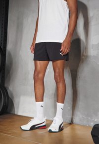 Svarta träningsshorts, vit linne, vita strumpor och Puma-sneakers med svarta, röda och blå detaljer. Slät textur och modern design.