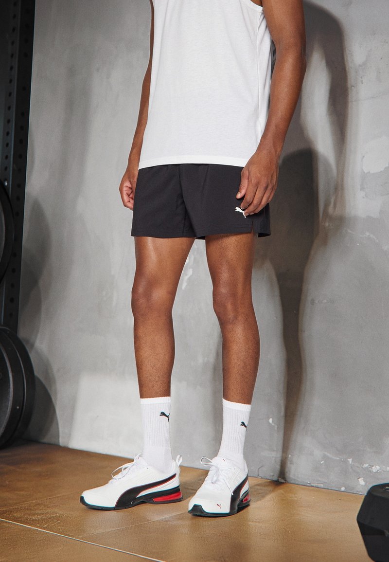 Svarta träningsshorts, vit linne, vita strumpor och Puma-sneakers med svarta, röda och blå detaljer. Slät textur och modern design.