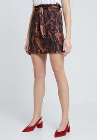 Hnědá mini sukně s hadím potiskem a páskem v pase, vyrobená z texturované látky. Doplněno červenými botami na blokovém podpatku pro kontrast.