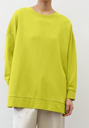 Personne portant un sweat-shirt ample vert citron à manches longues et un pantalon clair, debout devant un fond blanc uni.
