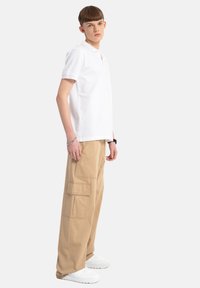 Biała koszulka polo z krótkim rękawem, zestawiona z beżowymi spodniami typu cargo z wieloma kieszeniami i białymi sneakersami. Prosty i casualowy design.