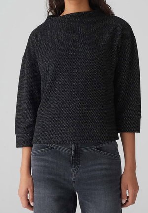 Vrouw die een zwarte, gestructureerde top met lange mouwen draagt, met een losse pasvorm en cropped donkergrijze jeans, staand tegen een lichtgrijze achtergrond.