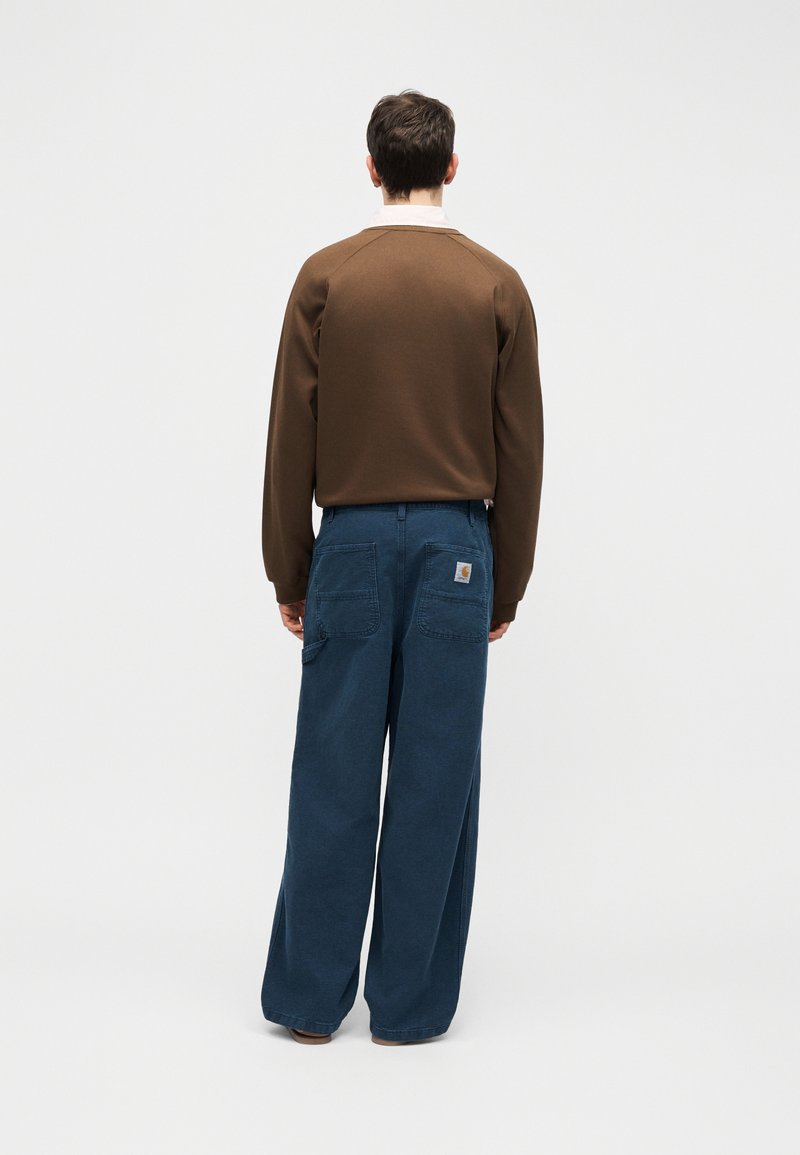 Homme portant une chemise marron à manches longues et un pantalon bleu ample, debout de dos contre un fond clair uni.