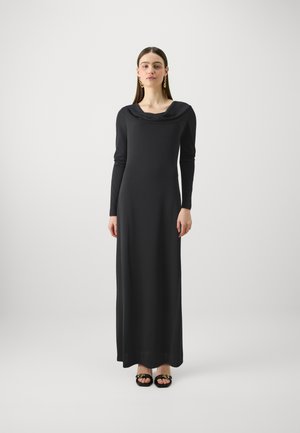 Pitkä musta mekko, jossa on kaulusnouto ja pitkät hihat, näyttäen istuvalta ja sileältä. Mustat sandaalit kultaisilla yksityiskohdilla.